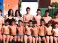 Estudiantes de una escuela secundaria posan en un patio deportivo, con uniformes escolares y bañadores. La imagen muestra a varios niños de diferentes edades, posando juntos en una fila, con un fondo que incluye un césped y una estructura de madera.
