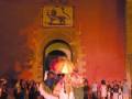 Una escena nocturna con un grupo de personas vestidas con trajes tradicionales, posiblemente en una celebración o evento cultural. En el fondo se puede ver un arco monumental con un león grabado en la puerta, típico de ciertas fortalezas históricas. La iluminación roja y el ambiente festivo sugieren una celebración importante, quizás un festival o una ceremonia tradicional.