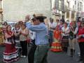 Un hombre en traje tradicional danza con una mujer en falda flamenca durante una celebración festiva. La imagen muestra a varias personas observando la danza, incluyendo una mujer en un vestido rojo y otro hombre con sombrero. La escena se desarrolla en una calle con edificios blancos y azules al fondo.