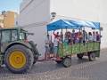 Una escena festiva con un tractor verde y azul, tirando de un remolque verde decorado. Un grupo de personas, incluyendo niños y adultos, se divierten en el remolque. El fondo muestra una calle con edificios blancos y un cielo claro.