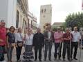 Una familia posa frente a una torre de la iglesia, con un hombre en el centro usando un sombrero y un bastón. La imagen muestra una escena típica de la vida en un pueblo español.