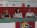 Imagen de una conferencia o evento con tres personas sentadas en un escenario. La imagen muestra un cartel que dice "XLVII ASCENSO DEL GUADALQUIVIR" y la fecha "13 DE OCTUBRE 2012". También hay una bandera de la "CLUB NAUTICO SEVILLA" en el fondo.