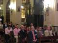 Una multitud de personas sentadas en bancos de madera, con vestimentas formales. Algunos llevan corbatas y otros no. En el fondo, una estructura de madera con inscripciones y un reloj en la parte superior. La iluminación es suave, proveniente de faroles y ventanas.