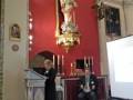 Un hombre y una mujer en un ambiente religioso, posiblemente una iglesia, frente a una imagen de Cristo en la cruz y un altar. La mujer está hablando o presentando algo, mientras el hombre escucha atentamente. En la pared hay un cuadro y una cruz de madera. La imagen se centra en el intercambio entre los dos personas y la atmósfera serena de la iglesia.