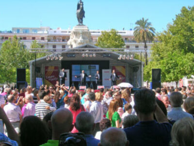 Una multitud de personas en un evento público, con una estatua en el centro y un escenario con luces en el fondo.