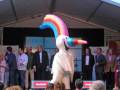 Una figura inflable de un unicornio con una lluvia de arcoíris en la cabeza se encuentra entre un grupo de personas en un escenario con una pancarta que dice "Fiesta". La iluminación es brillante y la atmósfera parece ser de una celebración o evento.
