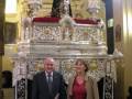 En una iglesia, un hombre y una mujer posan frente a una estatua religiosa en un altar dorado. La imagen transmite la solemnidad y el respeto hacia la religión.