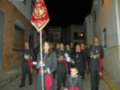 Un grupo de personas vestidas con uniformes militares caminan por una calle estrecha, seguidas de un niño que lleva un pequeño uniforme. La imagen muestra una celebración o desfile militar nocturno, con banderas y insignias visibles.