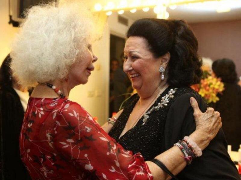 Dos mujeres mayores sonríen y abrazan, una con vestido rojo y la otra con negro brillante. La escena parece ser un evento formal, con luces de fondo y otros asistentes en el fondo.