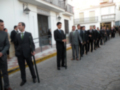 Una fila de hombres en trajes formales, algunos con armas, posiblemente en una ceremonia o evento oficial. La imagen muestra un entorno urbano con edificios blancos y una calle adoquinada.