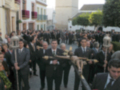 Una procesión religiosa en un entorno urbano, con personas vestidas formalmente y portando cruces de madera. La imagen muestra una torre en el fondo, junto a edificios históricos con balcones. La atmósfera es solemnemente festiva, probablemente durante una celebración religiosa o tradicional.