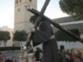 Una escena religiosa con un personaje vestido de negro sosteniendo una cruz, posiblemente en el contexto de la Semana Santa. La escena se desarrolla frente a un edificio con una torre, posiblemente una iglesia o templo. La gente observa la escena con atención, lo que sugiere una celebración o procesión religiosa.