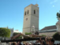 Una procesión religiosa con una cruz grande, personas en trajes formales y un edificio histórico con torre de ladrillo.