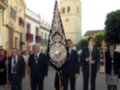 Un grupo de hombres vestidos formalmente, algunos con corbatas y otros con insignias, caminan por una calle adoquinada. Algunos llevan banderas y otros portan insignias. La arquitectura colonial de un edificio a la izquierda y una torre con dos campanas en el fondo completan el escenario.