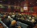 Una sala de conferencias con público sentado en filas. En el fondo, una pantalla proyecta imágenes y un panel de conferencia con personas hablando. La sala tiene paredes rojas y una gran pantalla en el centro.