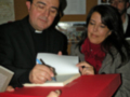 Un sacerdote firma un libro para una mujer, mientras un hombre observa en el fondo.