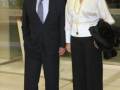 Una pareja posa en un entorno formal, el hombre viste un traje negro y la mujer una blusa blanca con pantalones negros. Detalles como un collar largo y una chaqueta de piel en la mano de la mujer son notables.