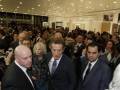 Una multitud de personas en trajes y chaquetas, posiblemente en un evento formal o ceremonial. La imagen muestra a varias personas de diferentes edades, incluyendo hombres y mujeres, algunas sonríen mientras otras parecen estar en silencio. El ambiente parece ser una sala con luces y decoración elegante.