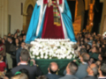 Una procesión religiosa en una iglesia, con una imagen venerada en un catafalco cubierto por flores blancas. La multitud observa la imagen con respeto y solemnidad, en un ambiente festivo.