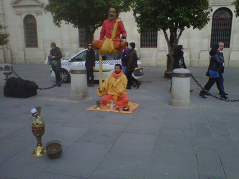 Una persona vestida de amarillo y rojo está sentada en el suelo, con un sombrero y una bolsa en el suelo a su lado, en una calle con personas y vehículos en el fondo.