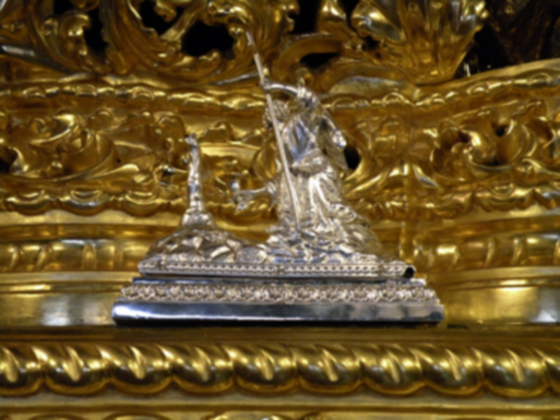 Una estatua de plata en relieve, posiblemente una figura religiosa o mitológica, sobre un pedestal dorado. El fondo es de oro con detalles en relieve que sugieren una escena histórica o mitológica.