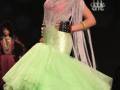 Una modelo presenta un vestido verde menta con detalles en rosa y negro, acompañado de una falda voluminosa. La imagen se realiza en el evento Fibes Sevilla, con un fondo negro y luces difuminadas.