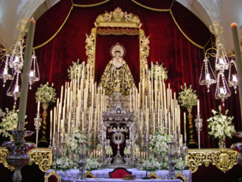 Altar con estatua central, velas altas y flores. Decoración dorada y roja. Lámparas de cristal.