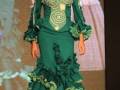 Modela en un desfile de moda flamenca con un vestido verde y detalles bordados, complementado con un tocado blanco y rojo.