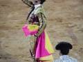 Un torero en traje tradicional de color rosa y amarillo, con una saya roja, levanta su sombrero negro mientras se prepara para la lucha. Detrás de él, otro torero con sombrero negro y traje azul se inclina para mirar al toro. La arena de la plaza de toros está vacía, enfocándose en el momento dramático del enfrentamiento.