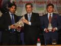 Premio Virgen de los Reyes. Tres hombres en trajes sostienen trofeos metálicos. Un hombre con corbata azul, otro con corbata roja y el tercero con corbata blanca. En la mesa hay una botella de agua y un vaso de cristal. Fondo con carteles informativos sobre el evento.