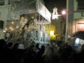 Fotografía nocturna de una procesión religiosa en una ciudad, con una imagen de la Virgen en el centro y una multitud de personas alrededor, iluminada por farolas y luces de la calle.