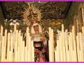 Una imagen de una Virgen con un manto rojo y blanco, rodeada por velas encendidas en una iglesia. La decoración dorada y el manto de la Virgen son destacables en esta escena religiosa.