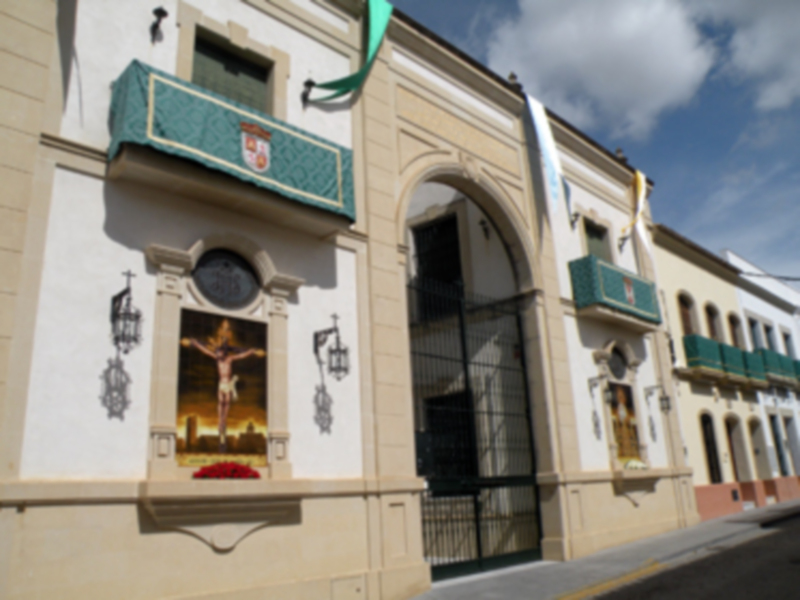 Fachada de un edificio histórico con una estatua del Cristo crucificado en la ventana. La fachada es de color beige con detalles en verde y dorado, y tiene un toldo azul con una bandera. La imagen muestra un cielo azul con algunas nubes.