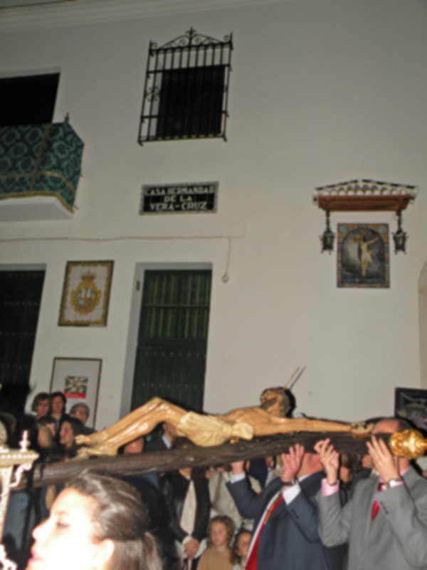Se observa una escena religiosa dentro de un edificio, probablemente una iglesia. En el centro se encuentra una cruz con la imagen de Cristo crucificado, sostenida por varias personas. La pared detrás muestra una ventana con reja y un cartel que dice "Casa Hermanada Vera Cruz". También se pueden ver otros carteles decorativos en la pared.
