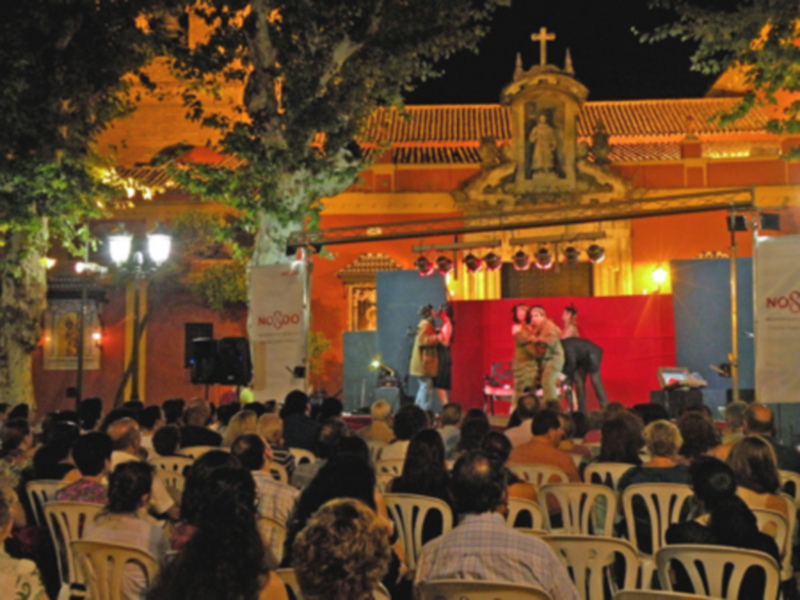 Escenario de teatro al aire libre con público en una noche de verano, iluminado por luces y decorado con banderines.