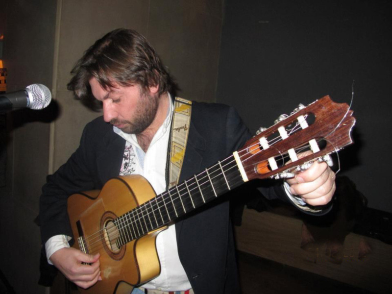 Un hombre con barba y cabello largo toca una guitarra clásica en un escenario. Detrás de él, se puede ver un micrófono y una pared de fondo oscuro.