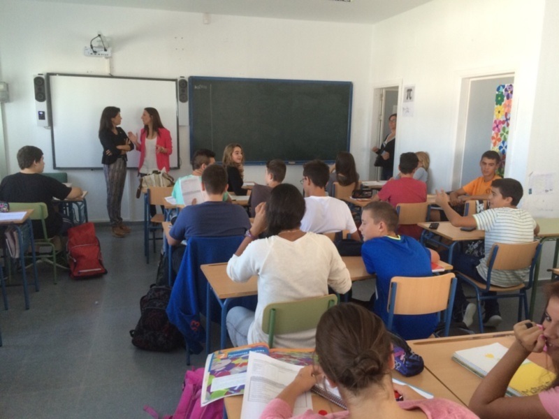 Clase de español en un aula con estudiantes sentados, una profesora hablando frente al pizarra y el estudiante en la mesa con un cuaderno abierto.