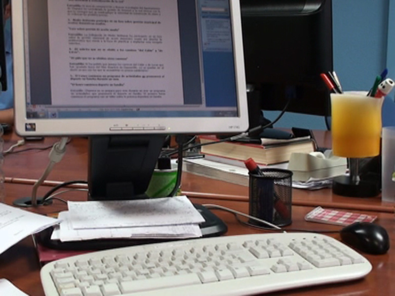 Un escritorio con un ordenador, una computadora portátil abierta mostrando texto en blanco y negro, un libro abierto, una taza de zumo, un marcador rojo en un portapapeles y un ratón.