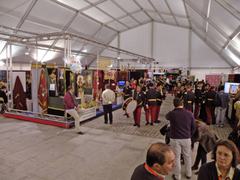 Una imagen de una feria o evento interior con personas en uniformes militares y civiles, disfrutando de stands y actividades. La estructura es una carpa blanca con iluminación artificial.