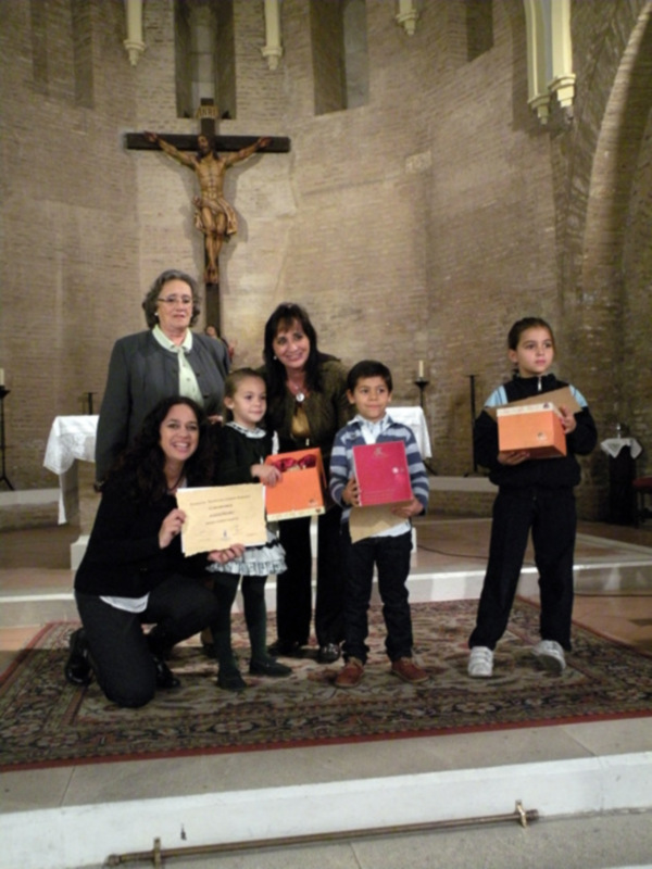 Una familia de cinco personas, incluyendo tres niñas y dos mujeres adultas, posan en una iglesia con un crucifijo en el fondo. Cada miembro de la familia sostiene una caja o un diploma, posiblemente como regalos o reconocimientos. La escena transmite una sensación de felicidad y orgullo familiar en un contexto religioso.
