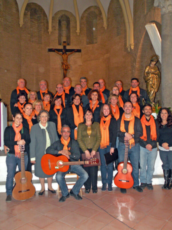 Un grupo de personas con bufandas naranjas posan en una iglesia, frente a un crucifijo y una estatua de la Virgen. Tres guitarras acústicas están a su lado.