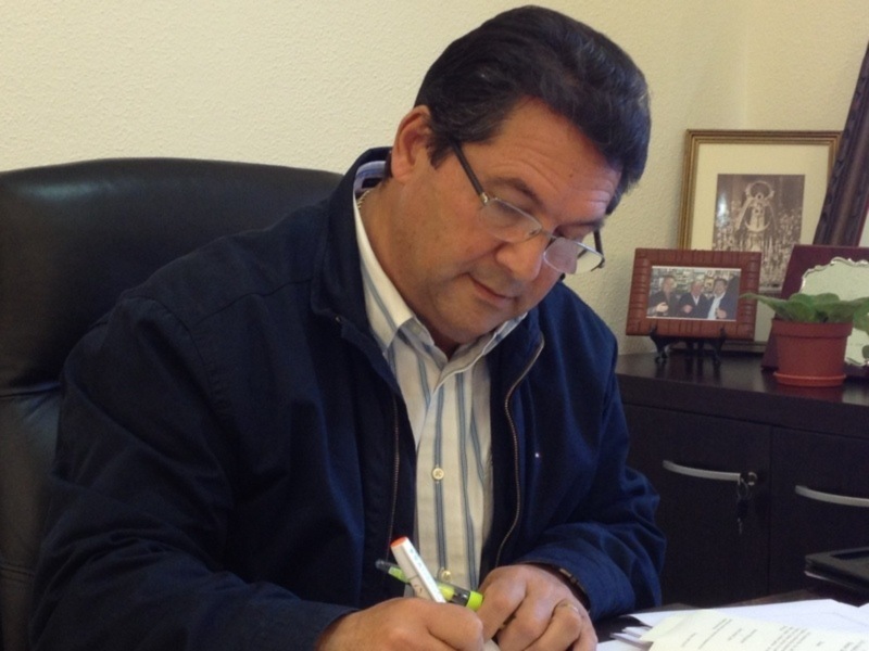 Un hombre está sentado en una silla de oficina, escribiendo en un papel con una pluma. Está vestido con una chaqueta azul y una camisa de rayas. En la mesa, hay un cuadro con fotos familiares y un maceta con una planta verde. El ambiente parece ser un lugar de trabajo o estudio.