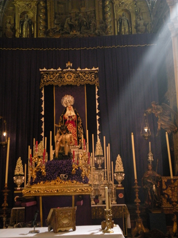Imagen de un altar religioso con una estatua central dorada y roja, rodeada por velas altas y candelabros. La atmósfera es solemnemente iluminada por un rayo de luz que cae desde arriba.