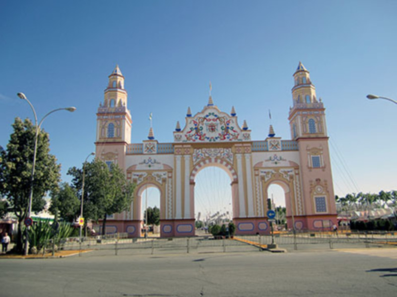 Puerta monumental con arcos y torres en un entorno urbano con árboles y cielo azul claro.