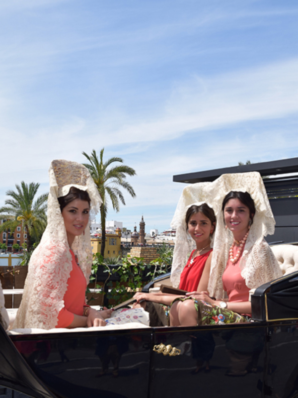 Festividad con mujeres vestidas con mantón de Manila en un carruaje, con palmeras y cielo azul al fondo.