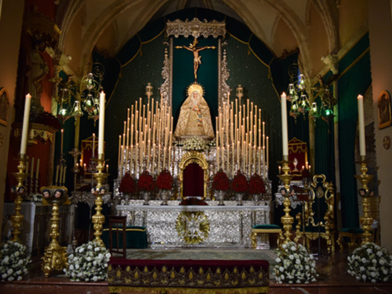 Altar de la iglesia con una Virgen en el centro, rodeada por velas y flores. La arquitectura es elegante con detalles dorados.