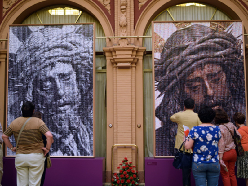 Dos grandes retratos de Jesús en mosaico colgados en una fachada con arcos y puertas de color beige. Personas observando los retratos mientras caminan por delante de la entrada principal. Detalles como una puerta con un cajón, una escalera y flores rojas en el suelo.
