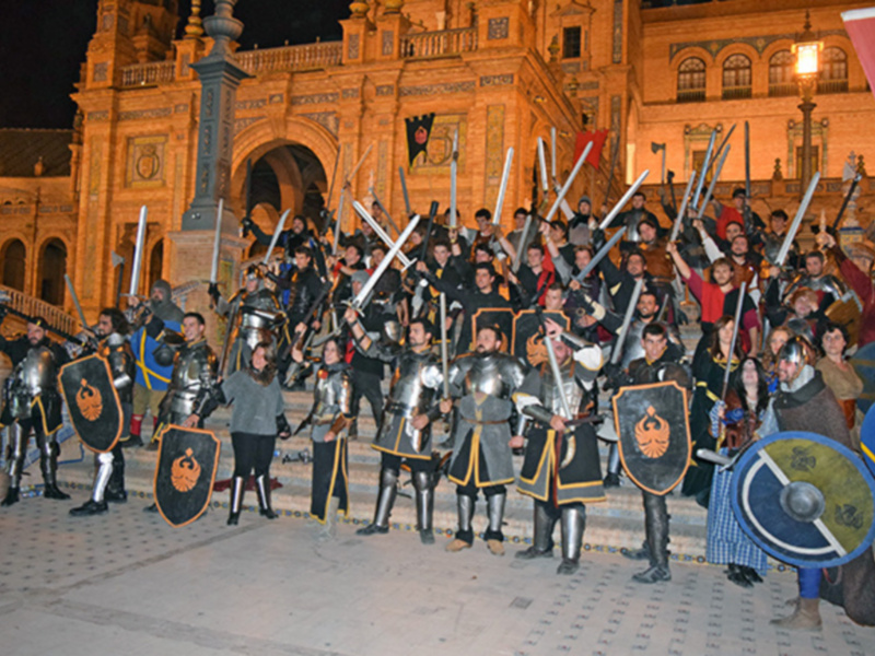 Un grupo de personajes vestidos con armaduras medievales y armas, posando en una plaza con un edificio histórico en el fondo, iluminado por faroles. La escena evoca una representación histórica o una recreación de la época medieval.