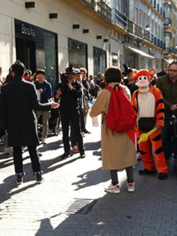 Un personaje disfrazado de Tigger camina por una calle con gente a su alrededor, en un día soleado en una ciudad europea. La calle está decorada con luces y los edificios son de estilo clásico.