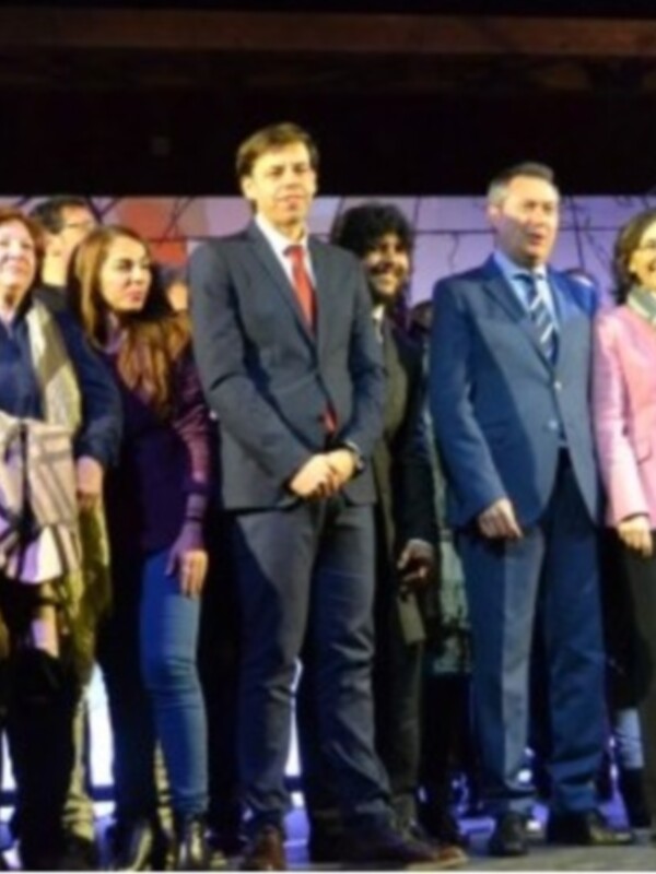Una imagen de una reunión formal en un escenario iluminado con luces de teatro. Una fila de personas, tanto hombres como mujeres, están posando para la foto. La mayoría llevan chaquetas y abrigos, indicando un clima frío. El fondo es una pantalla de color lila con luces que crean un ambiente festivo o ceremonial.